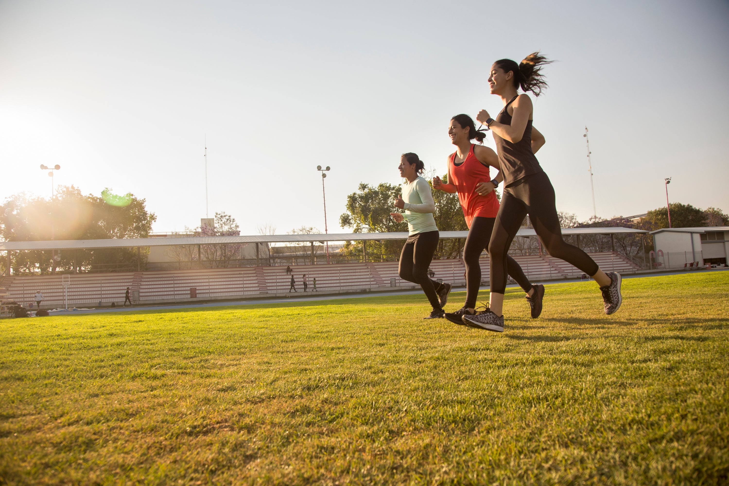 Three people running on a grassy field with stadium seats in the background