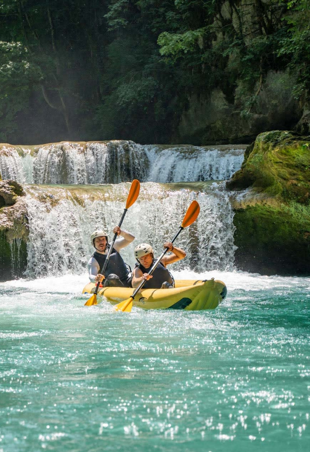 Two people kayaking in front of a waterfall with lush greenery.